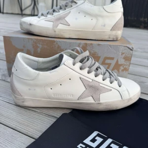 Golden Goose  - Golden Goose sneakers i vitt skinn med grå mockadetaljer och ikonisk stjärna på sidan. Skorna har grå snörning, platt sula och rund tå. Snyggt slitna detaljer på sulan för en cool, avslappnad look. Kommer med originalpåse och låda. Säljer då dem var lite små för mig, jag har 43 och storleken är lite mindre så passar perfekt på 42,5 och 42🌟 tveka inte att höra av dig vid fler frågor eller bilder🔥
