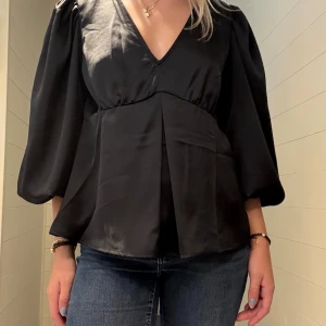 Svart satintopp  - Supersnygg svart topp i satin med djup v-ringning och vida puffärmar. Ärmarna har knytband vid handlederna. Är i nyskick utan prislapp och använd en gång. 