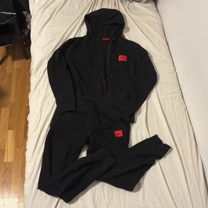 Svart hoodie och mjukis byxor från Hugo Boss - Svart hoodie från Hugo Boss med röd logotyp på bröstet. Tröjan har huva med snörning, dragkedja framtill och ribbade muddar. Tillverkad i mjuk bomull, perfekt för en chill och stilren look. Original pris för båda plagen är runt 2300 kr