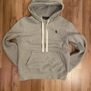 Grå hoodie från Polo Ralph Lauren - Snygg grå hoodie från Polo Ralph Lauren med klassisk broderad logga på bröstet. Tröjan har vit snörning i huvan, känguruficka och mjukt material som känns riktigt skönt. Perfekt för dig som gillar stilren och enkel streetwear.
