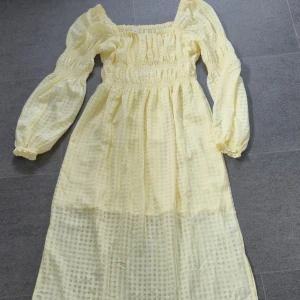 Ljusgul långklänning med puffärmar - Superfin ljusgul långklänning med rutigt mönster och puffiga långärmar. Klänningen har smock i midjan och vid ärmslut, samt fyrkantig ringning. Perfekt för dig som gillar romantiska och luftiga plagg med extra volym i ärmarna.