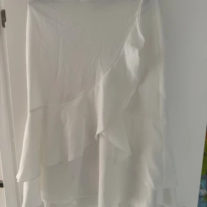 Vit omlottkjol med volang NA-KD - Superfin vit långkjol från NA-KD i omlottmodell med volangdetaljer. Kjolen har en flowy känsla och är tillverkad i ett glansigt, mjukt material som ger en lyxig look. Perfekt för dig som vill ha en stilren och trendig kjol med extra detaljer.