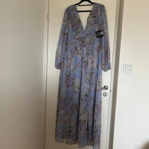 Ny blommig långklänning från NLY Eve - Säljer en ny drömmig långklänning från NLY eve  i ljusblått med blommigt mönster. Klänningen har v-ringning, långärmade ärmar och markerad midja. Modellen är ”remember me dress”. Dragkedja i ryggen som tyvärr har hoppat ur.