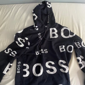 Svart hoodie från BOSS med tryck - Svart hoodie från BOSS med stora vita logotryck över hela tröjan. Tröjan har huva med snörning och långa ärmar. Materialet är mjuk bomull och passformen är normal. Perfekt för dig som gillar streetwear och tydliga loggor. Pris kan diskuteras 
