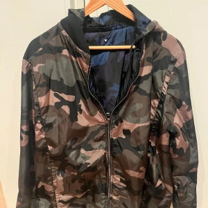 Camo valentino windbreaker - Säljer en snygg valentino windbreaker/vindjacka med huva och dragkedja, vändbar med två olika camouflage-mönster: ena sidan i grönt, brunt och svart, andra sidan i marinblått och svart. Storlek 50 motsvarande M/L. Mycket gott skick!