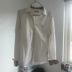 Vit Burberry Brit skjorta - Vit skjorta från Burberry Brit med klassisk krage och knappar framtill. Insidan av kragen och manschetterna har den ikoniska Burberry-rutiga detaljen i beige, svart och rött. Skjortan är långärmad och tillverkad i mjuk bomull. Perfekt för dig som gillar stilrena plagg med lyxig touch i ett väldigt bra skick för bra pris!