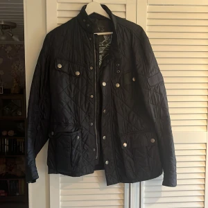 Svart quiltad jacka från Barbour - Quiltad svart jacka från Barbour International med ståkrage, tryckknappar och dragkedja. Jackan har flera fickor med knappstängning och Barbour-logga på ärmen. Perfekt för dig som gillar stilren och klassisk look med snygga detaljer.