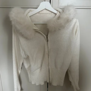 Vit hoodie med pälsdetalj på huvan - Säljer en vit hoodie med fluffig pälsdetalj på huvan och dragkedja framtill. Tröjan är stickad med ribbad nederkant och muddar vid ärmslut. Perfekt för dig som gillar mysiga och trendiga plagg med extra detaljer.