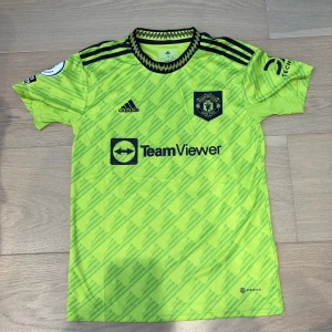Manchester United Garnacho bortatröja S - Manchester United bortatröja med Garnacho och nummer 49. Neon-gul med mörkblå detaljer, adidas-logga och klubbmärke på bröstet. Unikt mönster, svarta ränder på axlarna och Premier League-patch på ärmen. Tillverkad i lätt polyester med Aeroready-teknologi.