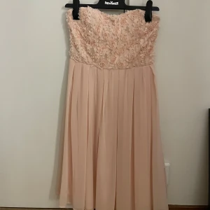 Ljusrosa axelbandslös klänning med rosor - Superfin ljusrosa axelbandslös klänning med 3D-blommor upptill och plisserad kjol. Överdelen har massor av små tygrosor som ger en romantisk vibe och kjolen faller snyggt i mjukt, lätt material. Perfekt för sommarens speciella tillfällen!