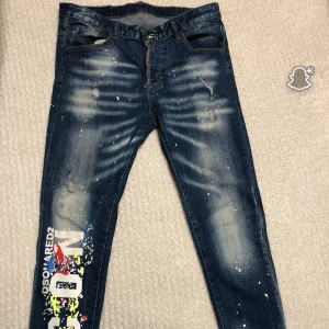DSQUARED2 jeans med färgstänk och tryck - Snygga blå jeans från DSQUARED2 med coolt färgstänk, slitningar och stort färgglatt tryck på ena benet. Klassisk femficksmodell med smal passform och tydliga slitningar för en edgy look. Tillverkade i bomull med detaljer i vitt, gult, rött och blått.