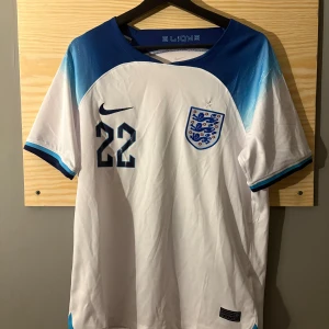 England hemmatröja VM 2022 - England fotbollströja med Bellingham och nummer 22. Vit bas med blå axelpartier och detaljer, officiellt klubbmärke på bröstet och Nike-logga. 