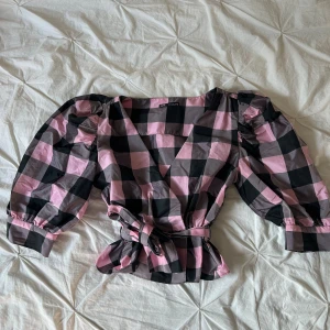 Rutig blus med puffärm från Zara - Superfin rutig blus från Zara i svart och rosa med stora puffärmar och v-ringning. Blusen har knytband i midjan och markerade axlar för en trendig siluett. Perfekt för dig som gillar statement-plagg och coola detaljer.