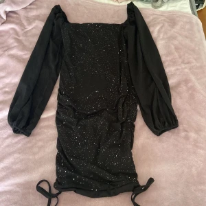 Svart glittrig kortklänning med långa ärmar - Svart kortklänning med glittriga detaljer och långa puffärmar. Klänningen har snörning nertill och dragkedja i ryggen. Tillverkad i stretchigt material med 40% polyester och 60% elastan. Perfekt för dig som vill sticka ut med en snygg och trendig look.