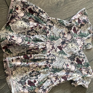 Blommig babyklänning från I Love Eco - Supersöt kort klänning från I Love Eco i ekologisk bomull. Klänningen har ett detaljerat blommigt mönster i ljusa och mörka toner med inslag av grönt, rött och beige. Korta ärmar med volang och tryckknappar vid axeln för enkel påklädning.