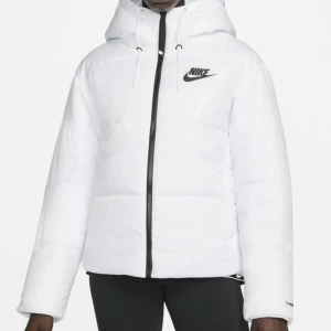 Vit jacka  - Snygg vit pufferjacka från Nike med svart logga på bröstet och dragkedja framtill. Jackan har huva med snörning och är vadderad för extra värme. På ärmen står det 'Therma-FIT'. Har bara använts några gånger och i väldigt bra skick, om ni har frågor är de bara och skriva. ( ber om ursäkt för dålig belysning)🥰