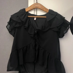 Svart volangblus från Nelly, strl 34 - Supersöt svart blus från Nelly med volanger längs ärmar, halsringning och nederkant. Blusen är gjord i ett lätt, transparent tyg och har korta ärmar. Perfekt för dig som gillar romantiska detaljer och flowy vibes.