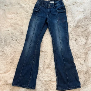 Blå bootcut jeans från Lindex - Snygga blå jeans från Lindex i bootcut-modell med coola fickor fram och bak. Byxorna har dekorativa knappar och ett broderat märke med rosor och texten 'True Love' baktill. Klassisk denim-look med markerade sömmar och lätt utsvängda ben.