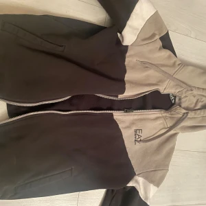 Svart och beige hoodie från EA7 - Snygg hoodie från EA7 Emporio Armani i svart och beige färgblock. Tröjan har dragkedja, huva med snörning och EA7-logga på bröstet. Perfekt för dig som gillar streetwear och vill ha en stilren look.