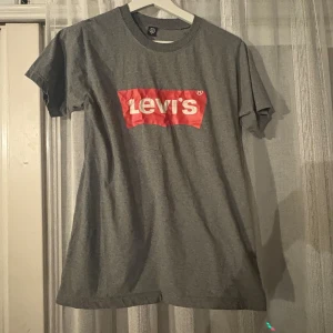 Grå Levi's t-shirt med röd logga - Snygg grå t-shirt från Levi's med klassisk röd logga framtill. T-shirten har rund halsringning och korta ärmar. Perfekt för en avslappnad och cool stil.
