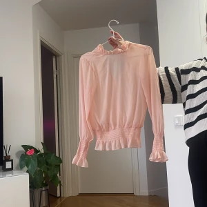 Ljusrosa blus med volangdetaljer - Supersöt ljusrosa blus med volang vid halsringning, ärmslut och midja. Blusen har långa ärmar och smockdetaljer vid handleder och midja som ger en snygg passform. Perfekt för dig som gillar romantiska och feminina plagg.