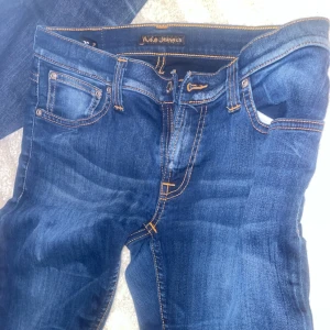  Nudie Jeans Co - Snygga blå jeans från Nudie Jeans Co med klassiska orange sömmar och ikonisk bakficka med vågig brodyr. Jeansen har fem fickor, normal passform och är tillverkade i slitstarkt denim. Perfekta för dig som gillar stilrena och tidlösa jeans.