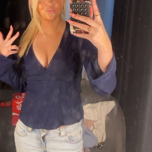 Mörkblå omlottopp med v-ringning - Säljer en mörkblå omlottopp med djup v-ringning och långa ärmar. Toppen har en figurnära passform och är tillverkad i ett mjukt, stickat material som känns skönt mot huden. Perfekt för dig som gillar en stilren men ändå trendig look.