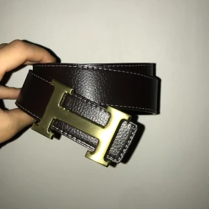  mörkbrunt skärp med H-spänne - Säljer ett mörkbrunt skärp i äkta skinn. Bältet har en stilren design med synliga vita sömmar och präglad storleksmärkning på insidan. Perfekt accessoar för att lyfta vilken outfit som helst.