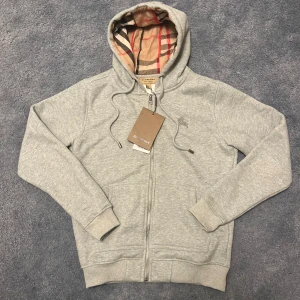 Grå hoodie från Burberry med dragkedja - Snygg grå hoodie från Burberry med klassiskt rutigt foder i huvan. Tröjan har dragkedja framtill, snörning i huvan och broderad logga på bröstet. Tillverkad i mjuk bomull med en skön passform. Perfekt för en chill och stilren look.