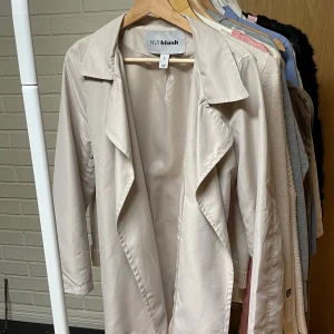 Beige trenchcoat från NLY Blush, S - Stilren beige trenchcoat från NLY Blush i storlek S. Jackan har klassisk krage, långa ärmar och bälte i midjan för snygg passform. Tillverkad i ett mjukt, lätt material som ger en clean look. Perfekt för dig som gillar minimalistisk och trendig stil.
