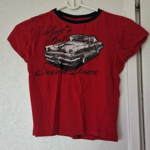 Röd t-shirt med biltryck - Röd t-shirt från Kappahl med svart/vitt vintage biltryck och texten 'Rock n Roll Tennessee Dream' på framsidan. T-shirten har svarta detaljer vid halsringningen och är i mjuk bomull. Perfekt för dig som gillar retro vibes. Trycket är sprucket och det är möjligt att färgen har mattats av något efter att ha tvättats.