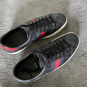 Gucci skor - Snygga svarta Gucci sneakers med klassiskt GG-mönster, röda och blå ränder på sidorna och röd detalj vid hälen. Skorna har snörning och vit platt sula. Materialet är skinn och textil, perfekt för dig som vill sticka ut med lyxig streetstyle.