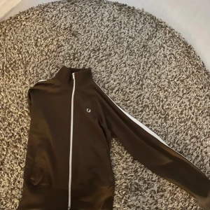 Fred perry - Detta är en brun track jacket med vita detaljer. Den har en dragkedja framtill, vita ränder längs ärmarna och ett litet Fred Perry-märke på bröstet. Designen är stilren och sportig.