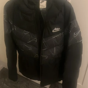 Svart pufferjacka från Nike L - Säljer en svart pufferjacka från Nike i storlek L. Jackan har ett fett mönster med vita Nike-loggor och linjer över bröst och rygg, samt en huva. Perfekt för kalla dagar och riktigt snygg streetstil.