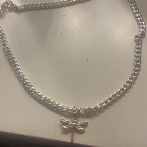 Wos halsband  - Chunky silverfärgat halsband med en söt trollsländeberlock. Kedjan är grov och glänsande, vilket ger ett coolt statement. Perfekt för dig som vill ha något unikt och trendigt runt halsen.