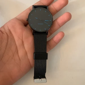 Svart minimalistisk klocka från Geneva - Stilren svart klocka från Geneva med mesh-armband och rund urtavla. Urtavlan är svart med tunna blå markeringar och visare, vilket ger en clean och modern look. Perfekt accessoar för dig som gillar enkel och snygg design.