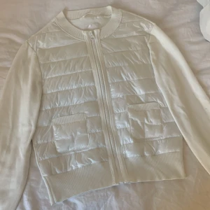 Vit quiltad cardigan jacka Gina Tricot - Snygg vit cardigan jacka från Gina Tricot med quiltad framsida och två fickor. Jackan har ribbade muddar, dragkedja framtill och är tillverkad i en mix av stickat och syntetmaterial. Perfekt för lager-på-lager och stilrena outfits.