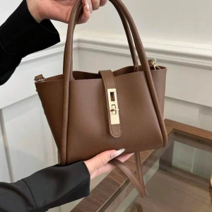 Brun handväska i skinn med gulddetalj - Snygg brun handväska i skinn med stilren design och guldfärgat spänne framtill. Väskan har två handtag och en axelrem, perfekt för att bära på olika sätt. Enkel men elegant look som passar till många outfits.