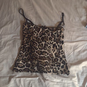 Leopardmönstrad linne-topp med smala band - Snygg linne-topp med leopardmönster i beige och svart. Tunt tyg med smala axelband och rak passform. Perfekt för dig som vill sticka ut med ett djurmönster. Toppen har en liten rosett framtill för extra detalj.