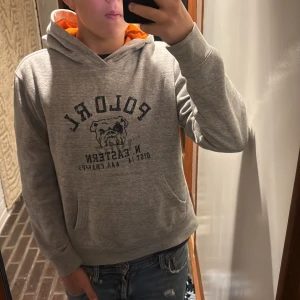 Grå hoodie Polo Ralph Lauren XL - Säljer en grå hoodie från Polo Ralph Lauren i storlek XL barn storlek. Tröjan har tryck med bulldog och text på bröstet, samt en orange insida i huvan. Klassisk känguruficka framtill och långa ärmar. Perfekt för dig som gillar sportig och avslappnad stil.