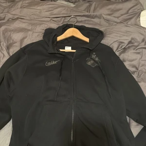 Svart AIK hoodie från Nike, storlek L - Svart hoodie från Nike med AIK-emblem och broderad Nike-logga på bröstet. Tröjan har huva med snörning, dragkedja framtill och en patch med texten 'Smoknglirare Edition 2025'. Perfekt för dig som vill representera AIK med stil. Tröjan är även använd max 5 gånger då jag köpte den online och tyckte den va för stor för mig som vanligtvis bär M/S. Båda snören är kvar. Ny pris 1200