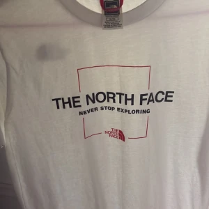 Vit t-shirt från The North Face XL - Snygg vit t-shirt från The North Face i bomull med tryck på både fram- och baksidan. Framsidan har logga och texten 'Never Stop Exploring' i svart och rött, medan baksidan har en mindre logga uppe vid axeln. Klassisk passform och rund hals.