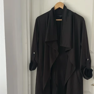 Svart lång trenchcoat med draperad krage - Säljer en svart lång trenchcoat med snyggt draperad krage och uppvikta ärmar med knappdetalj. Jackan har en loose passform och är tillverkad i ett mjukt, lätt material som faller snyggt. Perfekt för dig som gillar stilrena och minimalistiska plagg.