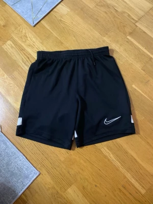 Svarta Nike shorts med vita detaljer - Snygga svarta shorts från Nike med vita detaljer på sidorna och bak. Shortsen har elastisk midja med snörning och är tillverkade i ett lätt och sportigt material som passar perfekt för träning. Nike-logga broderad framtill.