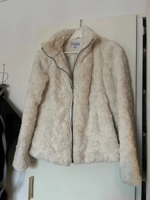 Vit pälsjacka från & Other Stories - Mysig creamvit pälsjacka från Stories med hög krage och dragkedja framtill. Jackan har långärmad design och är tillverkad i mjukt fuskpälsmaterial som ger en lyxig känsla. Perfekt för att hålla värmen under kyliga dagar.