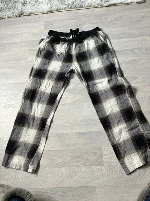 Rutiga pyjamasbyxor i svart och vitt - Säljer ett par sköna pyjamasbyxor med svartvitt rutmönster och dragsko i midjan. Byxorna har avslappnad passform och resår i midjan för extra komfort. Perfekta för mysiga hemmakvällar eller lata morgnar.