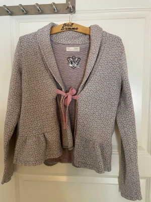 Mönstrad kofta från Odd Molly - Supersöt kofta från Odd Molly i ljusrosa och grått med diskret mönster. Koftan har volang nedtill, knyts med ett rosa band framtill.