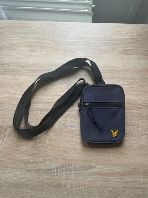 Mörkblå axelväska från Lyle & Scott - Mörkblå axelväska från Lyle & Scott med justerbar svart rem och två fack med dragkedja. Tillverkad i slitstark polyester och perfekt för att bära det viktigaste. Enkel, stilren och smidig design. Använd fåtal gånger.