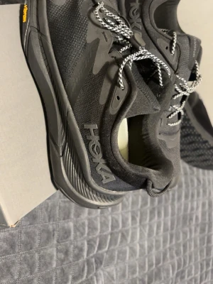 Svarta Hoka Transport sneakers - Snygga svarta Hoka Transport sneakers med reflekterande snören och robust sula. Ovandelen är i slitstark syntet och mesh för bra andningsförmåga. Hoka-logga på sidan och bekväm innersula. Perfekta för dig som gillar streetstyle och komfort.