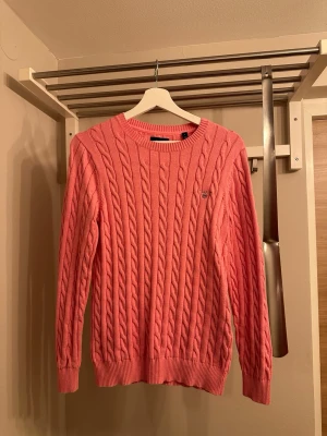 Rosa kabelstickad tröja från Gant - Snygg rosa kabelstickad tröja från Gant med rund halsringning och diskret logga broderad på bröstet. Tröjan har långa ärmar och ribbade muddar vid ärmslut och nederkant. Perfekt för dig som gillar klassisk och stilren design.